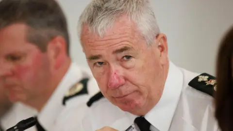 Chief Constable Jon Boutcher wearing a white shirt with psni shoulder titles. Behind him is another police officer in a white shirt. 