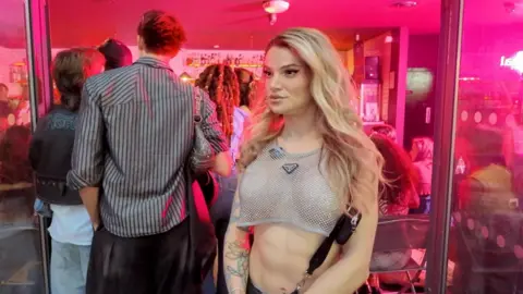 A woman with long, blonde hair wearing a mesh crop top looks at someone off-camera.