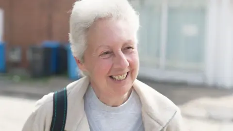 A smiling woman with grey hair stands in front of bins, wearing a grey t-shirt and cream top. She has a black satchel strap on her shoulder.
