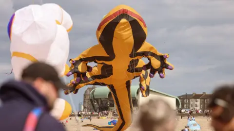 Redcar & Cleveland Borough Council/Dave Charnley A large yellow salamander kite with purple patches on the tips of its fingers. Blurred people can be seen in the foreground.