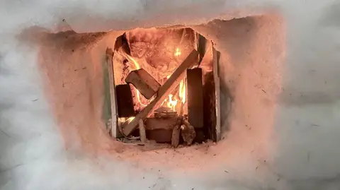 Kyle Doran Inside an igloo with a small fire burning in a wall niche, creating a warm glow against the snow interior.
