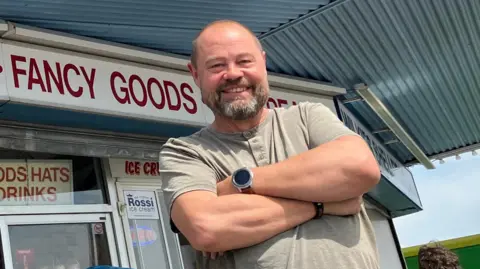 Henry Godfrey-Evans/BBC Chris Payne wearing a green top with his arms folded. He is smiling in front of his shop, which sells confectionary and souvenirs.