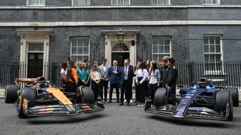 Reuters Two Formula 1 cars either side of the prime minster and several people looking at him and listening. The black door and grey brick of Number 10 Downing street is behind them