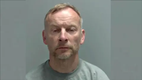 Norfolk Police A custody image shows Jonathan Creak, who has short, greying hair and stubble, looking towards the camera. He is wearing a grey sweatshirt and is in front of a plain grey background.