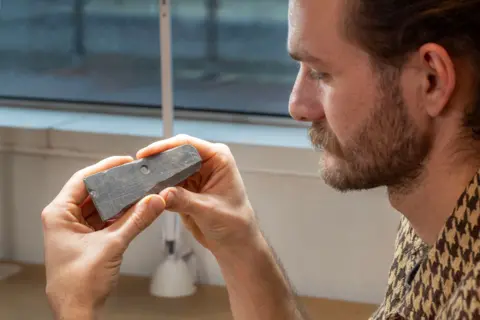 Mola/Andy Chopping A man with a beard and wearing a patterned shirt is looking at a piece of slate which has carvings on it