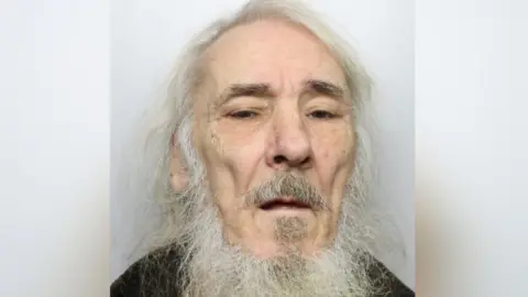 An elderly man with long white hair and beard. He is standing against a grey background wearing a black top.