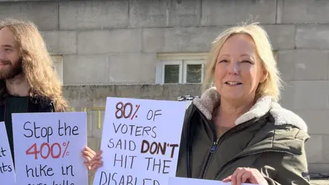 Chrissie Reidy/BBC A man with a beard and a blonde lady hold protest signs which read "stop the 400% hike in care bills" and "80& of voters said don't hit the disabled"