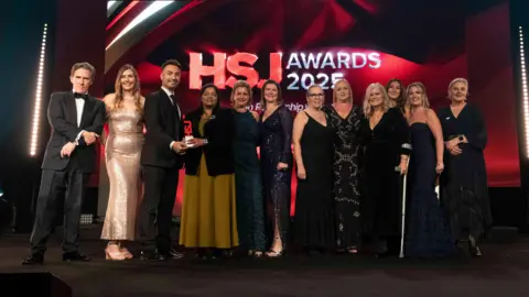 Health Service Journal A group of men and women stood on stage accepting an award, dressed in black tie