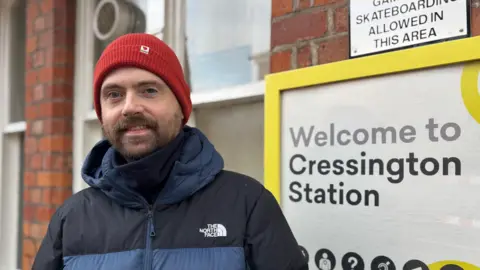 BBC John Mullany, with a brown beard and wearing a red hat and blue and black coat, stands in front of a sign saying Welcome to Cressington Station. He is vaguely smiling.