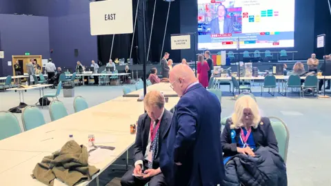 Shariqua Ahmed/BBC Paul Bristow and Wayne Fitzgerald at the Peterborough City Council election count, looking at a smartphone