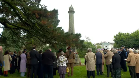 A large group of people in both civilian and military rig stand with their backs to the camera. The are stood outside at Victoria Gardens in Saltash. The group faces a large stone war memorial which has poppies laid at the base. An overhanging tree spans across the left of the image in the foreground. The weather is overcast.