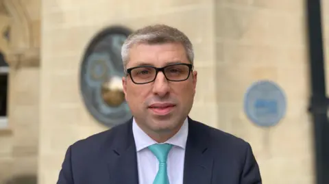 Mark Arnull with short dark hair and black-framed glasses wearing a light-coloured shirt, green tie and dark jacket. He is looking at the camera and is standing in front of the light stone wall of Northampton Guildhall