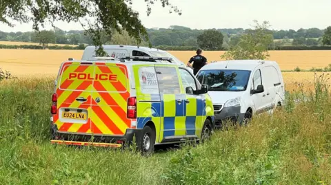 Three police vans parked on a field, with an officer looking away into the field.