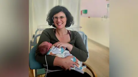 Layla Moran MP Layla sits on a hospital chair cradling a newborn baby. Layla has short dark hair and wears hexagonal glasses and a dark green long sleeve top. The baby is in a blue patterned onesie.