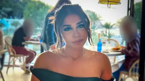 Ravika sitting in a restaurant, pictured head and shoulders looking directly at the camera. She has her hair tied up and is wearing a black off-the-shoulder dress.