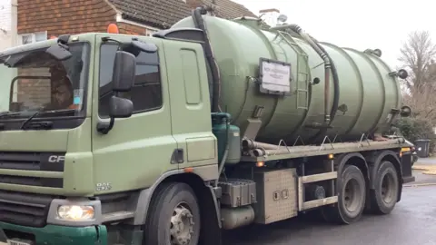 Nita Brown A green tanker parked outside homes. There are trees and bins in the background.