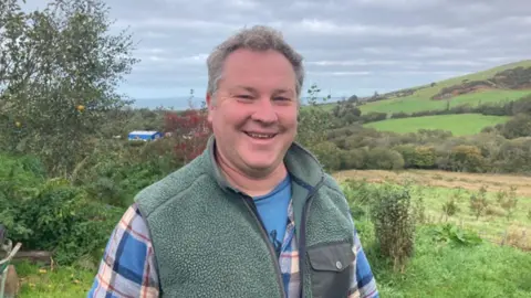 A middle aged man with curly grey hair, wearing a blue check shirt and a fluffy green gillet. He is stood in a field with more grassy fields and hills behind him and is smiling as he looks at the camera.