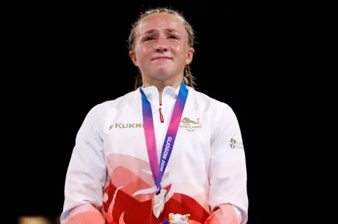 Reuters Ruby White wearing a Team England tracksuit stands with a Glasgow 2026 medal around the neck against a dark background.