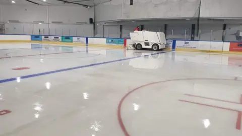 An ice rink is empty with barriers around the side with adverts on. You can see a person driving a white ice resurface vehicle around the edge of the rink.