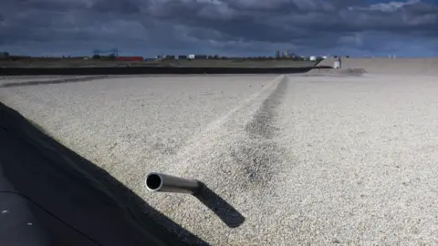 Augean A newly engineered landfill cell with a silver pipe emerging from the gravel. The industrial landscape can be seen in the background.