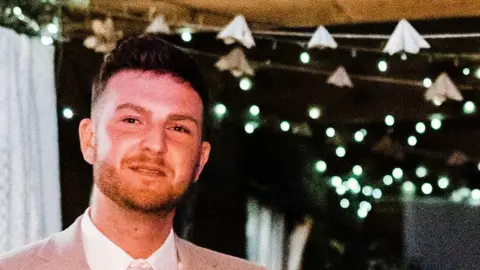 Tom  A young white man with short black hair. He's wearing a white shirt and cream suit jacket. Behind him we can see strings of fairy lights and white, sheer fabric hanging from a gazebo or awning of some sort. It suggests he's at a wedding.