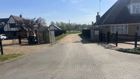 Two silver gates are in between two houses. Both of them are open and a driveway leads to a field. There is a 10mph sign on a pillar on the right and a defibrillator is on the left gate.