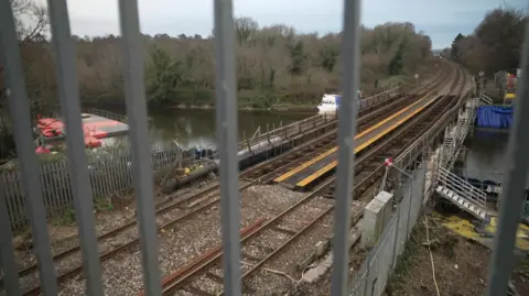 A railway bridge over a river, seen through the beams of a metal fence. There are trees and some buildings on either side of the tracks beyond the bridge. There is a floating plastic platform in the water to the left of the bridge.