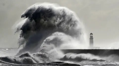 PA Media A huge wave rises from the sea, crashing into a harbour. There is a lighthouse to the right which is dwarfed by the wave.