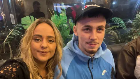 Kevin Lacey A photo of a woman with curly blonde hair pictured next to a boy with a light blue tracksuit. They both have little smiles. He is wearing a cap and looking up. 