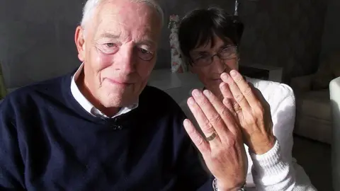 BBC / Simon Spark Christopher Brightmore has receding grey hair and blue eyes. He is wearing a blue jumper and is sitting next to his wife, who is wearing a white jumper and metal, oval-shaped spectacles on her face. They are both holding their hands up to the camera showing their wedding rings, which they are wearing on their ring finger.