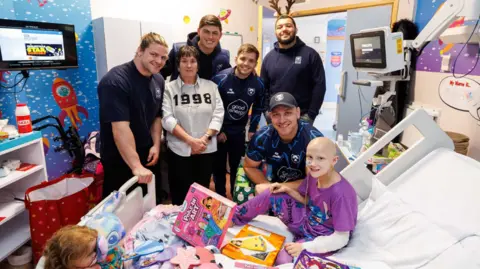Bristol Bears Several Bristol Bears players and a woman stand by the hospital bedside of a child aged about 10 in purple pyjamas surrounded by toys. The child has no hair and a tube is coming out of her nose. A hospital monitor is visible behind her head.