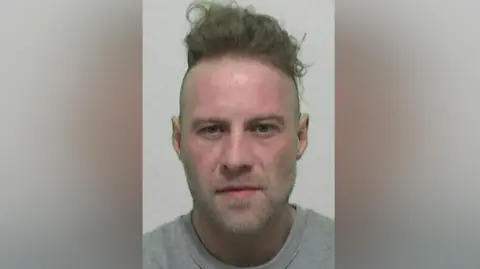 Northumbria Police Mugshot of Henry. He has long brown hair stacked on top of his head with shaved sides and slight stubble around his chin.