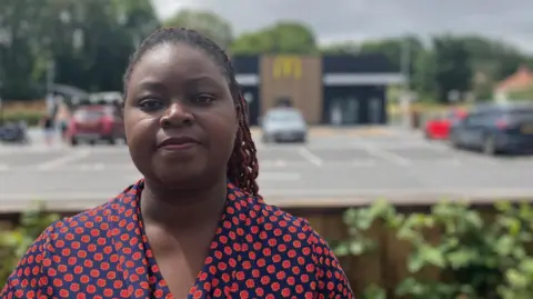 Makala Zulu looks towards the camera wearing a red and blue patterned top, with a McDonald's restaurant blurred in the background