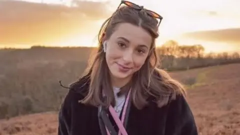 Debbie Hollinson Darcy Hollinson, who has long brown hair and sunglasses on her head, looks toward the camera smiling
