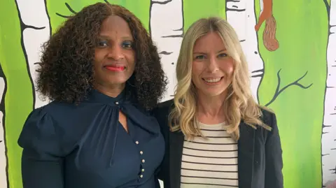 A woman with shoulder-length black curls is wearing a navy blue blouse and is standing next to a woman with shoulder-length blonde curls who is wearing a striped top and dark blue blazer. They are stood in front of a wall which is painted bright green with white tree trunks and a squirrel.