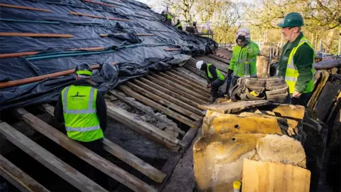 Historic Property Restoration Ltd Four construction workers on the barn roof. The rafters are exposed at the bottom with a man sitting on them, and black plastic protects the higher rafters. Three other men in high vis and hard hats watch while standing by piles of yellow insulation