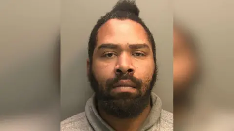 Police custody photograph of a head and shoulders shot of Joshua Springer. He has black hair and a beard and is wearing a grey hoody and pulling a serious expression. 