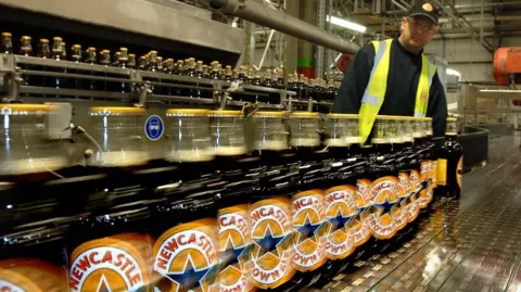 PA Media A plant worker inspects one of the first bottles of Newcastle Brown Ale as it comes off the bottling line at the Federation Brewery, Gateshead, in May 2005. The bottles contain a dark liquid while the label says "Newcastle Brown Ale". There is a blue star in the centre of the logo.