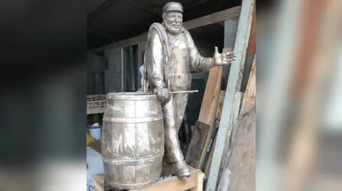 A metal cast of a statue showing a standing man dressed like a fisherman, wearing a cap and with his left arm and hand out, as if gesturing while in conversation. His right hand is on an upright barrel to his right hand side. 