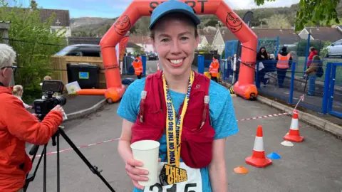 A female runner smiles at the camera at the finish line of the race. She is wearing a blue baseball cap, a blue t-shirt and a red running vest. She has a yellow "The Big Cheese" medal around her neck and holds a cup in one hand