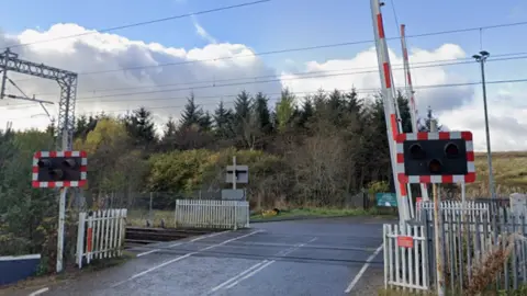 Google A train crossing on a road. The barrier is lifted and the lights on either side are off