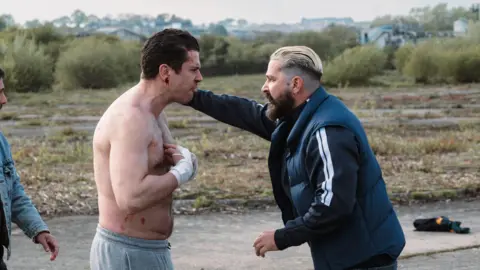 Lionsgate Shia LaBeouf and Toby Kebell stand angrily looking at eachother face to face on a yard. Labeouf stands on the right hand side with his hand on Kebell's shoulder. has bleached blonde hair slicked back and cut short on the sides. He wears an earring and has a dark brown long beard. He wears a black zip up top with two white stripes on the sleeves and a black gilet. Kebell is topless with grey joggers on, he has blood coming from his ear and his hands are bandaged.