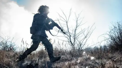 Getty Images A soldier in uniform and helmet advancing across a battlefield with his gun at the ready on his shoulder. The landscape looks wintry. There appears to be frost on the ground and trees and shrubs have no leaves. The soldier is partly silhouetted by the sun, which is shining directly at the camera.