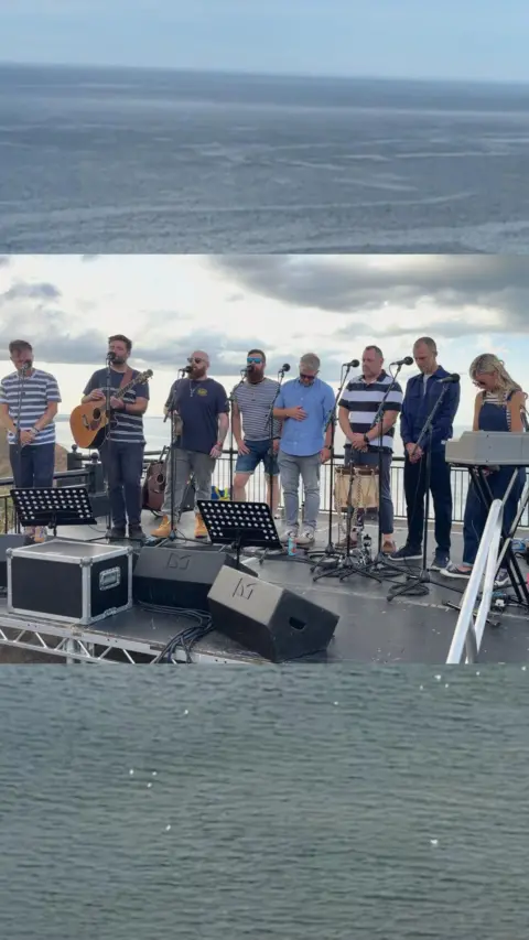 A group of eight performers stand on a stage outside singing into microphones. The view of the sea is behind them. One performer holds an acoustic guitar while another stands at a keyboard. 