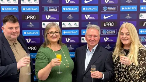 Lee Sanders, Frances Connolly, Patrick Connolly and Dawn Hepple standing in a row holding flutes of champagne. Mrs Connolly's glass has orange juice in it instead. Mr Sanders has dark, short hair and is wearing a blue suit jacket. Mrs Connolly has shoulder-length grey hair and is wearing a green long-sleeved top with a long, gold necklace. Mr Connolly has short, grey hair and is wearing a blue suit jacket and lighter blue shirt. Ms Hepple has long, blonde hair and is wearing an animal print blouse.