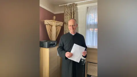 Supplied The Reverend Johannes Arens is standing holding a letter with a statue of Christ in the background.