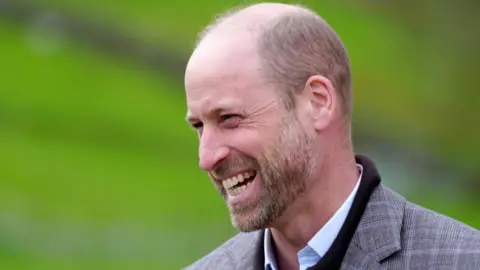 Prince William smiling. He is dressed in a tweed jacket worn over a dark jumper and light blue shirt. 
