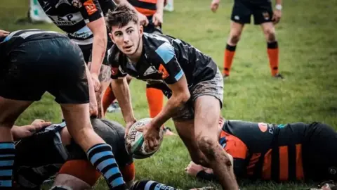 Family Handout Freddie Pring is seen playing rugby with his teammates. He is wearing his black rugby kit and is holding the ball.