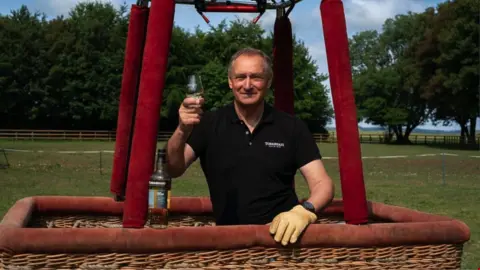 PA Media Sir David Hempleman-Adams standing in the basket of a hot air balloon holding a glass