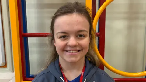 A head-and-shoulders shot of Maisie Summers-Newton smiling directly at the camera. Her hair is half up, half down. She is standing in front of a yellow and red climbing frame. She is wearing a GB Paralympic blue zip-up hoodie.